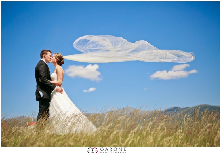Bride and groom kiss in a field with a flowing veil under a blue sky.