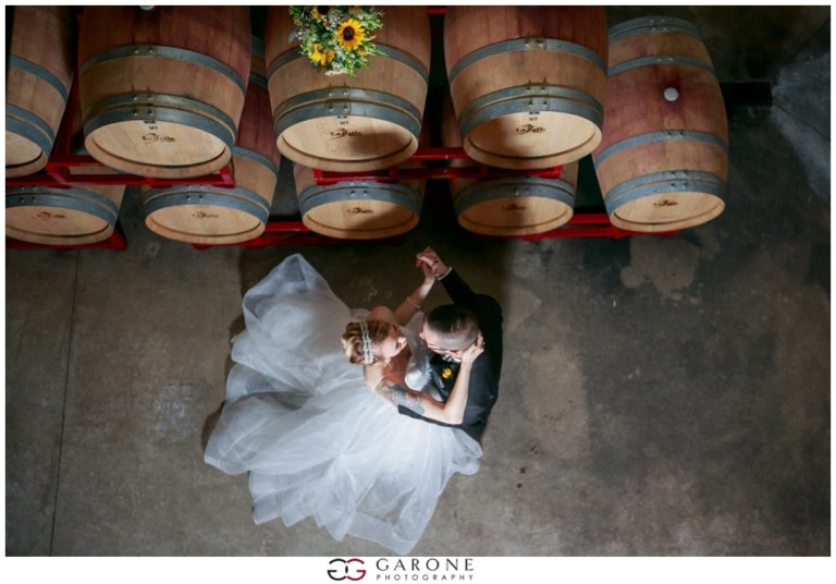 Bride and groom dancing beneath wine barrels in a rustic cellar.