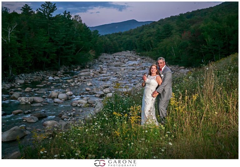 Newlyweds posing by a rocky river with forest and mountains behind.