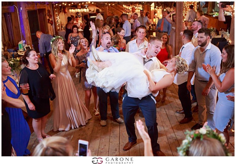 Groom lifting bride joyfully during wedding celebration surrounded by guests.