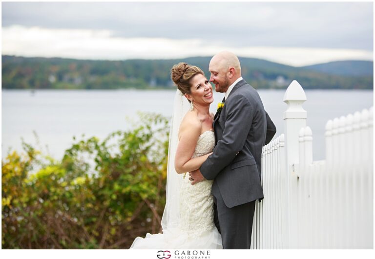 Bride and groom smiling by lakeside fence.