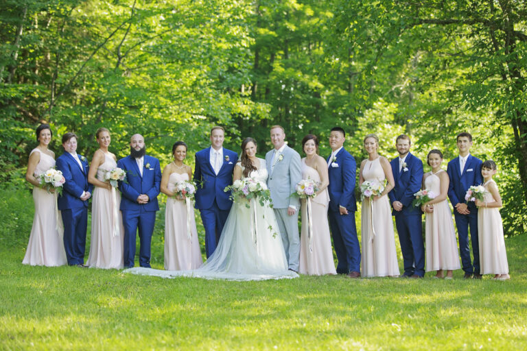 A wedding party posing outdoors in formal attire under lush green trees.