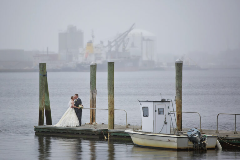 A couple embraces on a foggy pier with boats and industrial cranes in the background.