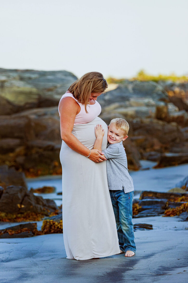 A pregnant woman in a white dress embraces a young boy by the rocky shore.